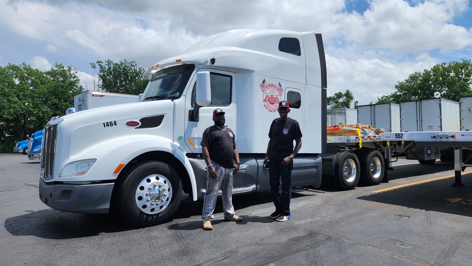 Delta team drivers Jack Drew and Jamie Gray in Delta gear beside their white Peterbilt 579 with a loaded flatbed, unit 1464
